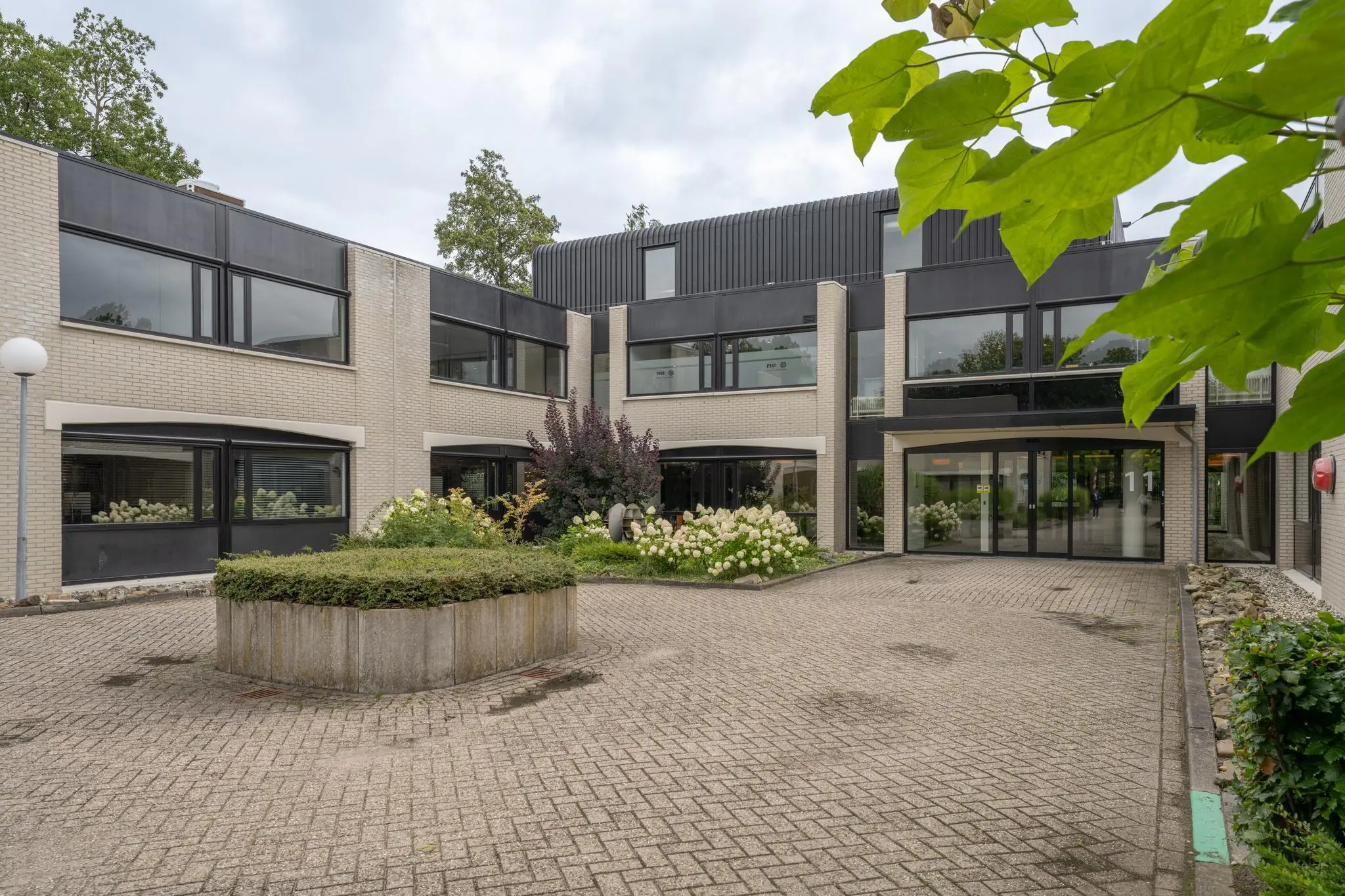 Ingang van een modern kantoorgebouw aan de Burgemeester Verderlaan met een betegelde binnenplaats en bloeiende planten.